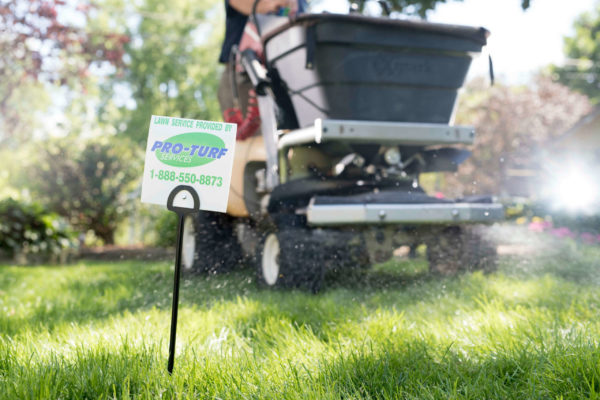A PRO-TURF Services lawn care professional applies fertilizer with an Ex Mark applicator, walking by a "Lawn Service Provided by PRO-TURF Services" sign.